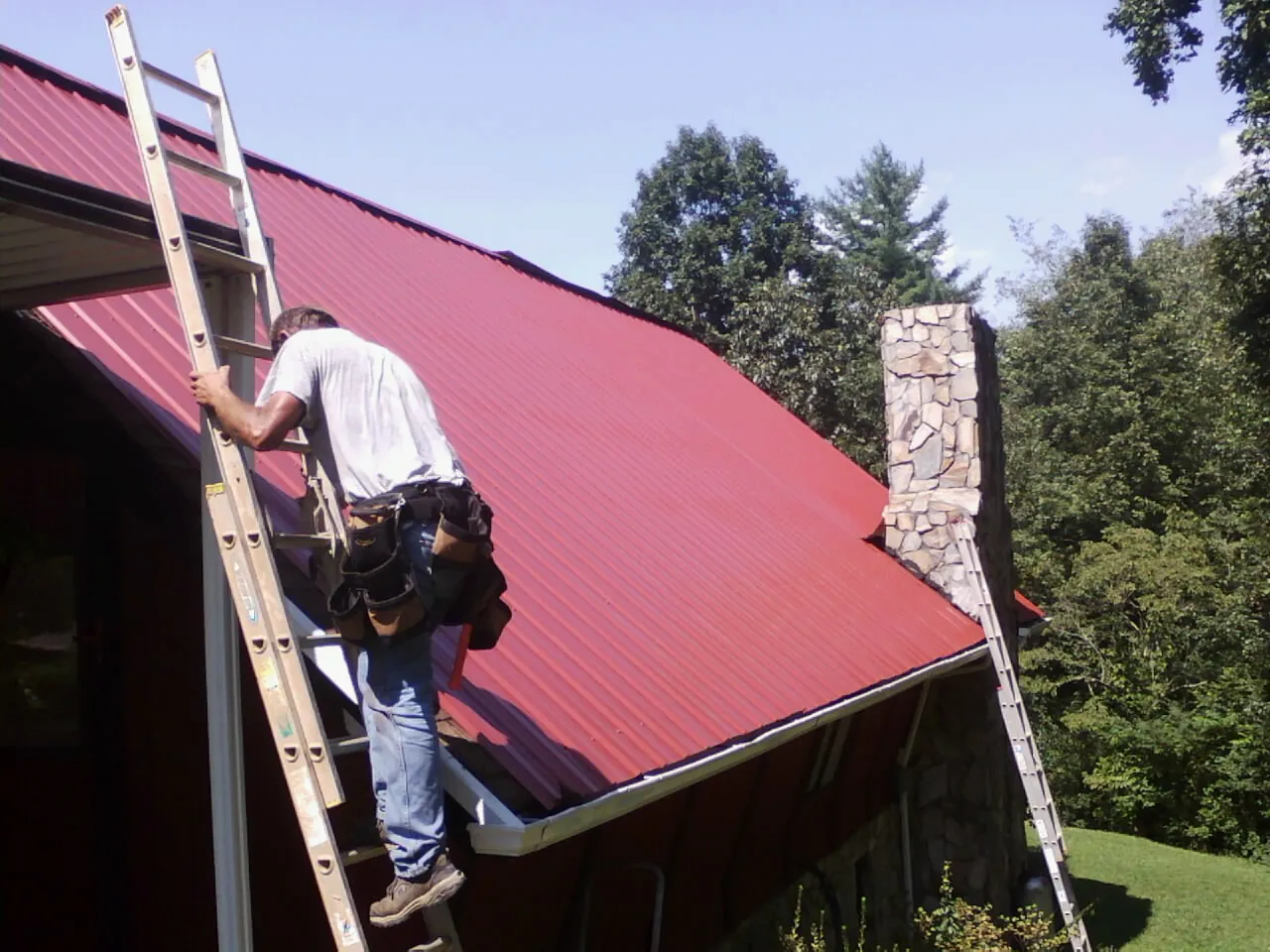 Roofer on a ladder beside a red metal roof and stone chimney — Soffit & Fascia Repair by Roofing Brothers in Goose Creek